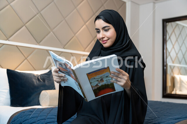 مفهوم الثقافة والتعلم الذاتي, الاستمتاع بالأنشطة الترفيهية المفيدة في وقت الفراغ, قراءة القصص والروايات, الإقامة في إحدى الفنادق الفاخرة، امراة عربية خليجية اماراتية ترتدي العباءة السوداء تجلس على سرير في غرفة النوم تحمل بيدها كتاب بإيماءات البشاشة والفرح