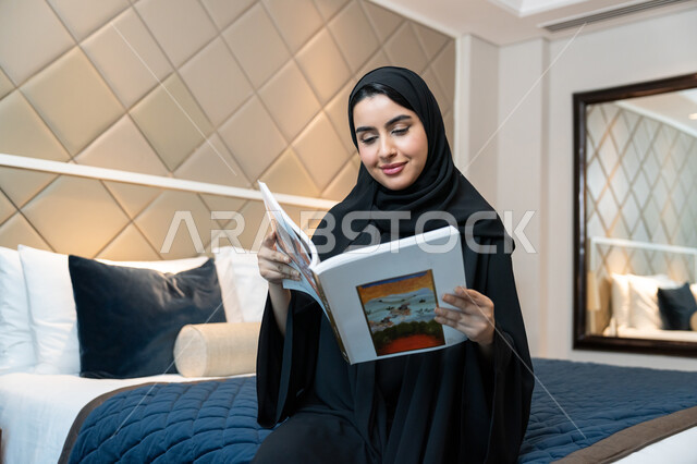 Staying in one of the Kingdom's luxurious hotels, the concept of culture and self-learning, enjoying useful recreational activities in free time, reading stories and novels, an Emirati Gulf Arab woman wearing a black abaya sitting on a bed in the bedroom holding a book in her hand with gestures of cheerfulness and joy