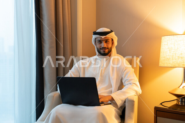 Looking at the camera with gestures of pleasure and cheerfulness, using modern advanced technologies, managing and organizing business affairs, holding a business meeting online, portrait of a young Arab Gulf Emirati man wearing a white kandura and ghutra sitting on a comfortable sofa working on a laptop
