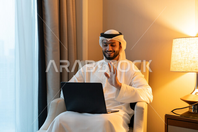 إجراء مكالمة فيديو من خلال الحاسب المحمول, ايماءات السرور و البشاشة, عقد إجتماع عمل عبر الإنترنت، استخدام التقنيات الحديثة المتطورة، بورتريه لشاب عربي خليجي اماراتي يرتدي الكندورة والغترة البيضاء يجلس على اريكة مريحة يقوم برفع يده بتعابير القاء التحية والترحيب