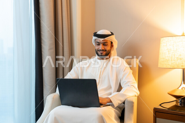 ايماءات السرور و البشاشة, عقد إجتماع عمل عبر الإنترنت، استخدام التقنيات الحديثة المتطورة، إدارة شؤون الأعمال وتنظيمها، بورتريه لشاب عربي خليجي اماراتي يرتدي الكندورة والغترة البيضاء يجلس على اريكة مريحة يعمل عبر الحاسب المحمول