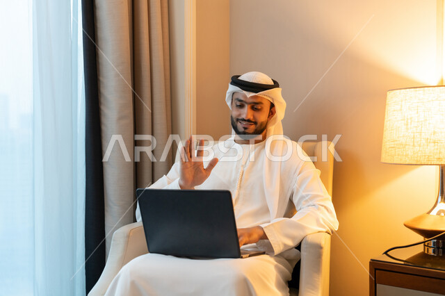 إجراء مكالمة فيديو من خلال الحاسب المحمول, ايماءات السرور و البشاشة, عقد إجتماع عمل عبر الإنترنت، استخدام التقنيات الحديثة المتطورة، بورتريه لشاب عربي خليجي اماراتي يرتدي الكندورة والغترة البيضاء يجلس على اريكة مريحة يقوم برفع يده بتعابير القاء التحية والترحيب