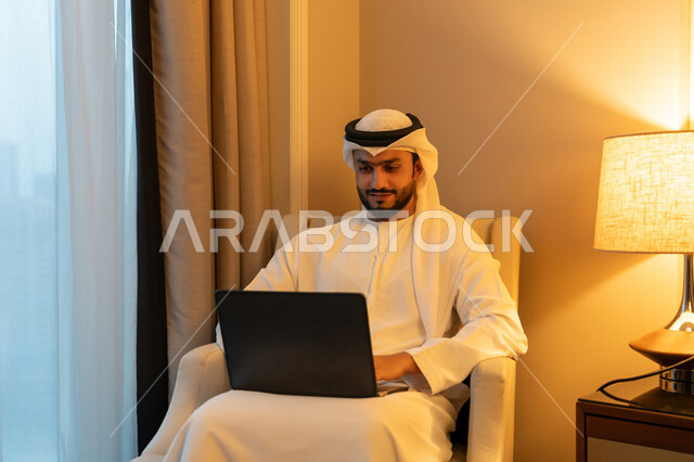 Using advanced modern technologies, holding a business meeting online, managing and organizing business affairs, portrait of a young Emirati Gulf Arab man wearing a white kandura and ghutra sitting on a comfortable sofa working on a laptop, gestures of pleasure and Cheerfulness
