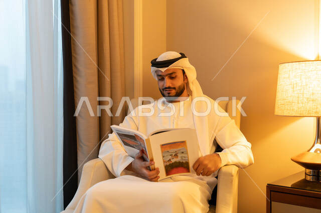 قراءة القصص والروايات, الاستمتاع بالأنشطة الترفيهية المفيدة في وقت الفراغ, مفهوم الثقافة والتعلم الذاتي, ممارسة هواية المطالعة, بورتريه لشاب عربي خليجي اماراتي يرتدي الكندورة والغترة البيضاء يجلس على اريكة مريحة يحمل بيده كتاب بإيماءات البشاشة والفرح