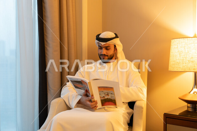 الاستمتاع بالأنشطة الترفيهية المفيدة في وقت الفراغ, مفهوم الثقافة والتعلم الذاتي, قراءة القصص والروايات, ممارسة هواية المطالعة, شاب عربي خليجي اماراتي يرتدي الكندورة والغترة البيضاء يجلس على اريكة مريحة يحمل بيده كتاب بإيماءات البشاشة والفرح