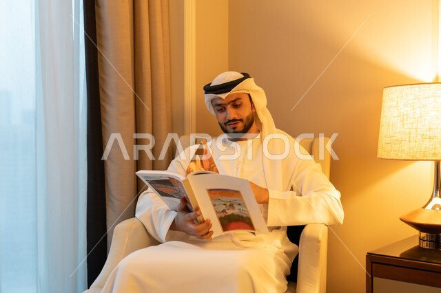 The concept of culture and self-learning, enjoying useful recreational activities in free time, reading stories and novels, practicing the hobby of reading, a portrait of a young Arab Gulf Emirati man wearing a white kandura and ghutra sitting on a comfortable sofa holding a book in his hand with gestures of cheerfulness and joy