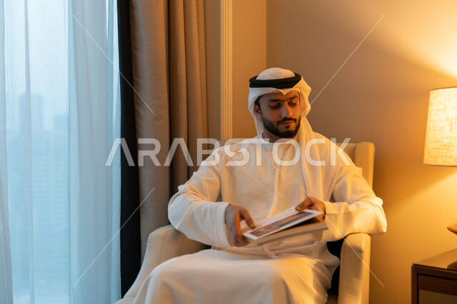 ممارسة هواية المطالعة, مفهوم الثقافة والتعلم الذاتي, الاستمتاع بالأنشطة الترفيهية المفيدة في وقت الفراغ, قراءة القصص والروايات, شاب عربي خليجي اماراتي يرتدي الكندورة والغترة البيضاء يجلس على اريكة مريحة يحمل بيده كتاب بإيماءات الجدية و الحزم