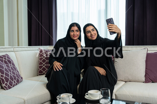 توثيق اللحظات السعيدة باستخدام الهاتف المحمول, تعابير الاستمتاع و الفرح, امراة عربية خليجية اماراتية محجبة ترتدي العباءة السوداء تجلس على اريكة مريحة تقوم بالتقاط صورة سيلفي مع شقيقتها, العلاقة القوية والترابط مع الأصدقاء, زياره عائليه في غرفة المعيشه