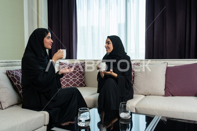 ايماءات السعادة و الفرح, الاستمتاع بالمشروبات الساخنة, مفهوم التحدث مع الأصدقاء ومناقشة الأخبار، امراة عربية خليجية اماراتية محجبة ترتدي العباءة السوداء تجلس على اريكة مريحة تقوم باحتساء القهوة مع شقيقتها, الكرم وحسن الضيافة