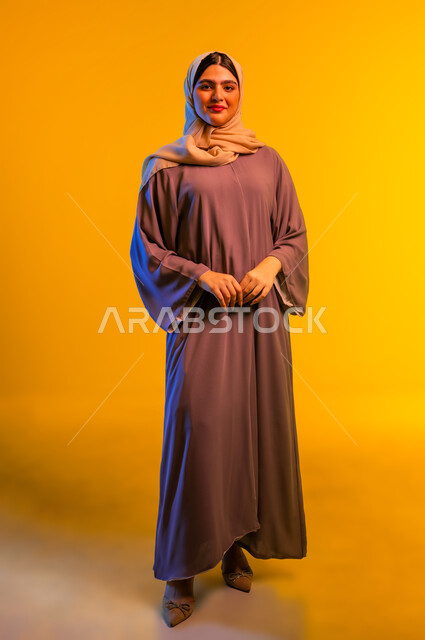 سيدة سعودية تنظر إلى الكاميرا بإيماءات السرور والسعادة، الجمال والاهتمام بالمظهر الخارجي، مفهوم الأنوثة والنعومة، بورتريه لامرأة عربية خليجية عمانية ترتدي الحجاب والعباءة تقف باستقامة بتعابير الثقة بالنفس، الجسم بالطول الكامل، خلفية صفراء