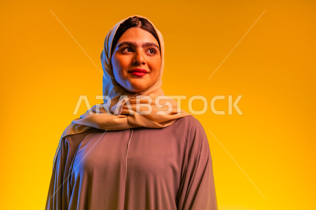 Concept of femininity and softness, close-up portrait of an Arab Gulf Omani woman wearing hijab and abaya looking at something with gestures of pleasure and happiness, Saudi lady watching something above, beauty and interest in outward appearance, yellow background