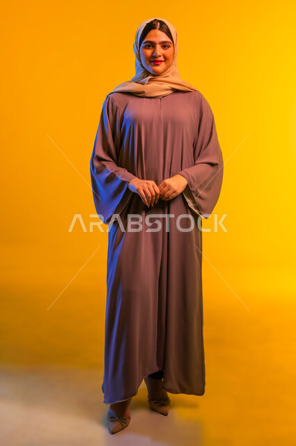 الجمال والاهتمام بالمظهر الخارجي، مفهوم الأنوثة والنعومة، بورتريه لامرأة عربية خليجية عمانية ترتدي الحجاب والعباءة تقف باستقامة بتعابير الثقة بالنفس، سيدة سعودية تنظر إلى الكاميرا بإيماءات السرور والسعادة، الجسم بالطول الكامل، خلفية صفراء