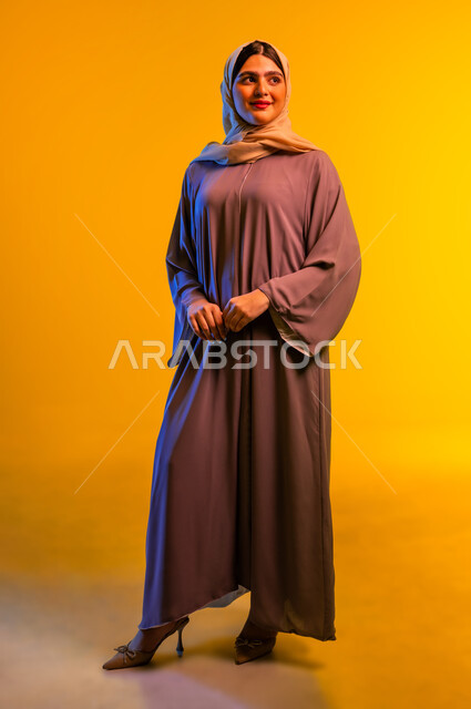 Concept of femininity and softness, portrait of an Arab Gulf Omani woman wearing hijab and abaya standing straight with self-confident expressions, Saudi lady looking at something with gestures of pleasure and happiness, beauty and interest in outward appearance, full length body, yellow background