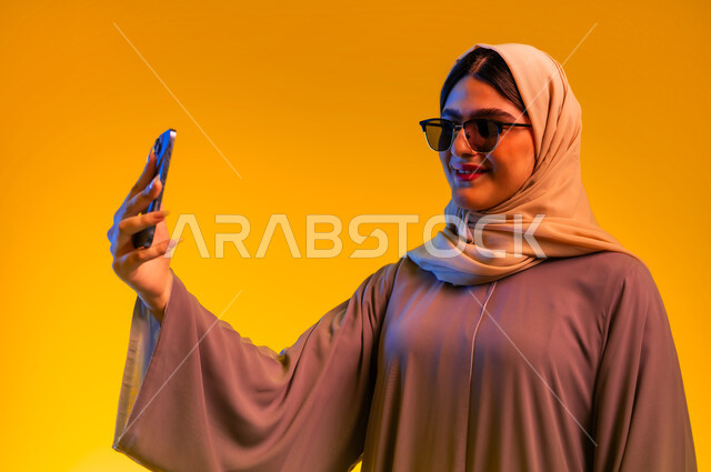 أجهزة تقنية حديثة، سيدة سعودية تقوم بالتقاط صور سيلفي، إجراء مكالمة فيديو مع الأهل والأصدقاء، الاستمتاع بمتابعة البرامج الترفيهية، بورتريه مقرب لشابة عربية خليجية عمانية ترتدي العباءة الملونة والحجاب والنظارات الشمسية تستخدم تطبيقات الهاتف المحمول، خلفية صفراء