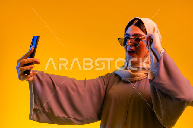 استخدام التكنولوجيا والأجهزة التقنية الحديثة، إيماءات التفاجؤ والذهول، سيدة سعودية تقوم بالتقاط صور السيلفي, بورتريه مقرب لشابة عربية خليجية عمانية ترتدي العباءة الملونة والحجاب والنظارات الشمسية تقوم بإجراء مكالمة فيديو عبر الهاتف المحمول, خلفية صفراء