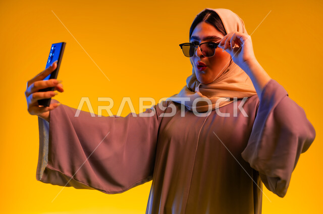 الاستغراب والانبهار والحماس, استخدام الأجهزة التقنية الحديثة، إيماءات التفاجؤ والذهول، سيدة سعودية تقوم بإجراء مكالمة فيديو, بورتريه مقرب لشابة عربية خليجية عمانية ترتدي العباءة والحجاب والنظارات الشمسية تستخدم تطبيقات الهاتف المحمول, خلفية صفراء