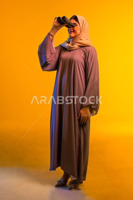 التفكير والتطلع إلى المستقبل، التخطيط المستقبلي للأهداف والطموحات، سيدة سعودية تقوم برؤية الأشياء البعيدة، حب الاستكشاف والمغامرة، بورتريه لشابة عربية خليجية عمانية ترتدي العباءة والحجاب تمسك منظار ثنائي العينين تنظر إلى شيء ما، الجسم بالطول الكامل، خلفية صفراء