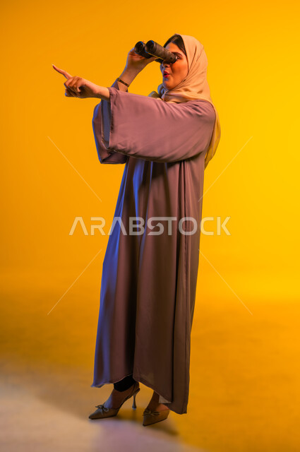 سيدة سعودية تقوم برؤية الأشياء البعيدة، حب الاستكشاف والمغامرة، بورتريه لشابة عربية خليجية عمانية ترتدي العباءة والحجاب تمسك منظار ثنائي العينين تشير بإصبع السبابة إلى شيء ما، التخطيط المستقبلي للأهداف والطموحات، الجسم بالطول الكامل، خلفية صفراء