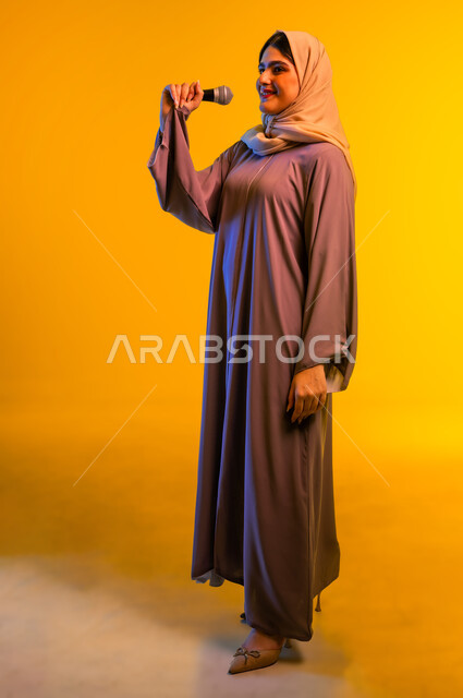تعابير الاندماج والتفاعل، أجهزة تقنية حديثة، بورترية لامرأة عربية خليجية عمانية ترتدي الحجاب والعباءة تضع سماعات الرأس تمسك بيدها مكبر الصوت تنظر إلى شيء ما، سيدة سعودية تقوم بإلقاء الخطابات والمحاضرات، ممارسة المواهب الفنية، الجسم بالطول الكامل، خلفية صفراء