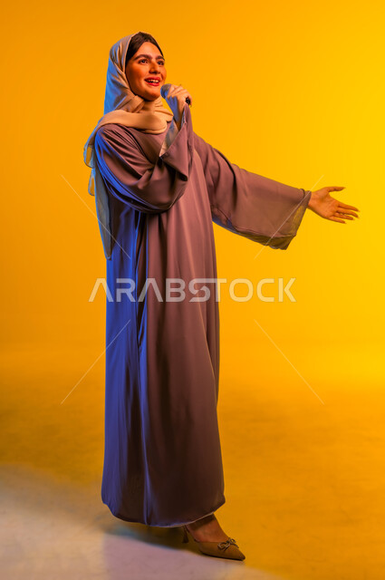 استخدام الأجهزة التقنية الحديثة، بورترية لامرأة عربية خليجية عمانية ترتدي الحجاب والعباءة تضع سماعات الرأس تمسك بيدها مكبر الصوت تنظر إلى شيء ما، سيدة سعودية تقوم بإلقاء الخطابات والمحاضرات، ممارسة المواهب الفنية، الجسم بالطول الكامل، خلفية صفراء