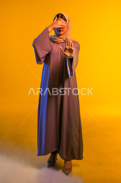الأمر بالتوقف عن فعل شيء ما، سيدة سعودية تحاول عدم رؤية حدث مزعج، بورتريه لشابة عربية خليجية عمانية ترتدي العباءة والحجاب تضع يدها على عيونها واليد الأخرى إلى الأمام بإيماءات النهي، الجسم بالطول الكامل، خلفية صفراء