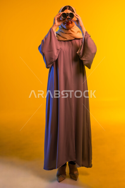 سيدة سعودية تقوم برؤية الأشياء البعيدة، حب الاستكشاف والمغامرة، بورتريه لشابة عربية خليجية عمانية ترتدي العباءة والحجاب تمسك منظار ثنائي العينين، التخطيط المستقبلي للأهداف والطموحات، التفكير والتطلع إلى المستقبل، الجسم بالطول الكامل، خلفية صفراء