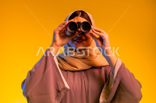 التخطيط المستقبلي للأهداف والطموحات، سيدة سعودية تقوم برؤية الأشياء البعيدة، حب الاستكشاف والمغامرة، بورتريه مقرب لشابة عربية خليجية عمانية ترتدي العباءة والحجاب تمسك منظار ثنائي العينين تنظر إلى شيء ما، التفكير والتطلع إلى المستقبل، خلفية صفراء