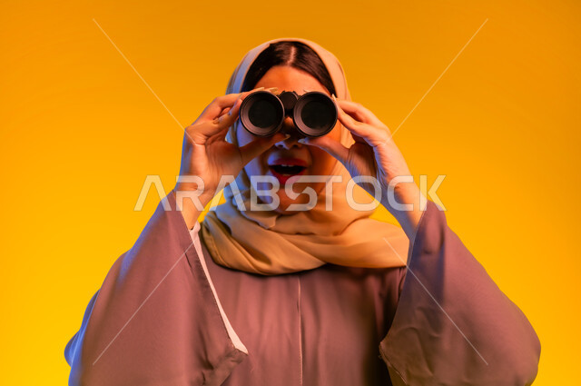 التخطيط المستقبلي للأهداف والطموحات، سيدة سعودية تقوم برؤية الأشياء البعيدة، حب الاستكشاف والمغامرة، بورتريه مقرب لشابة عربية خليجية عمانية ترتدي العباءة والحجاب تمسك منظار ثنائي العينين تنظر إلى شيء ما، التفكير والتطلع إلى المستقبل، خلفية صفراء