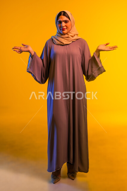 التساؤل والاستغراب والاستفسار من أمر ما، بورتريه لامرأة عربية خليجية عمانية ترتدي الحجاب والعباءة تنظر إلى الكاميرا تفتح ذراعيها بإيماءات عدم المعرفة، سيدة سعودية تقوم بالاستفهام حول موضوع معين, الغموض وعدم الوضوح، الجسم بالطول الكامل، خلفية صفراء