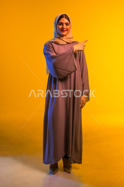 سيدة سعودية تقوم بتحديد موقع معين بإيماءات البشاشة، بورتريه مقرب لامرأة عربية خليجية عمانية مبتسمة ترتدي الحجاب والعباءة ترفع إصبعها السبابة تشير إلى شيء ما، إشارات وحركات يدين تدل على هذا المكان (هنا)، الجسم بالطول الكامل، خلفية صفراء