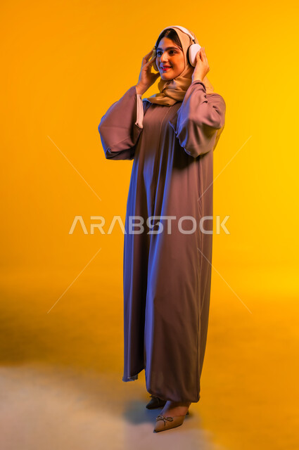 أجهزة تقنية حديثة، سيدة سعودية تقضي أوقات ممتعة بسماع الأغاني، بورتريه لامرأة عربية خليجية عمانية ترتدي الحجاب والعباءة تضع سماعات الرأس تنظر إلى شيء ما، الاندماج مع الأنغام الموسيقية، مفهوم الإصغاء واستخدام تقنيات الهيدفون، الجسم بالطول الكامل، خلفية صفراء