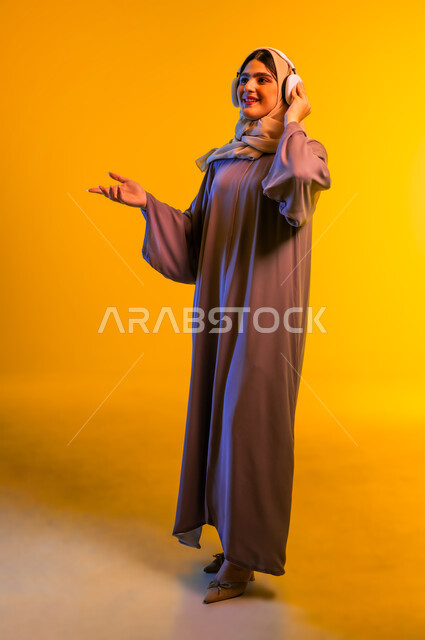 مفهوم الإصغاء واستخدام تقنيات الهيدفون، أجهزة تقنية حديثة، سيدة سعودية تقضي أوقات ممتعة بسماع الأغاني، بورتريه لامرأة عربية خليجية عمانية ترتدي الحجاب والعباءة تضع سماعات الرأس تنظر إلى شيء ما، الاندماج مع الأنغام الموسيقية، الجسم بالطول الكامل، خلفية صفراء