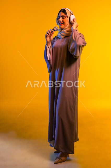 ممارسة المواهب الفنية، أجهزة تقنية حديثة، بورتريه لامرأة عربية خليجية عمانية ترتدي الحجاب والعباءة تضع سماعات الرأس تمسك بيدها مكبر الصوت، سيدة سعودية تقوم بإلقاء الخطابات والمحاضرات، مفهوم الإصغاء واستخدام تقنيات الهيدفون، الجسم بالطول الكامل، خلفية صفراء