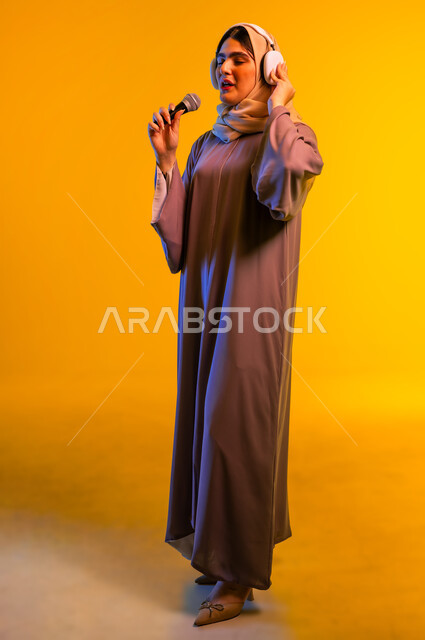 أجهزة تقنية حديثة، بورتريه لامرأة عربية خليجية عمانية ترتدي الحجاب والعباءة تضع سماعات الرأس تمسك بيدها مكبر الصوت، سيدة سعودية تقوم بإلقاء الخطابات والمحاضرات، ممارسة المواهب الفنية، مفهوم الإصغاء واستخدام تقنيات الهيدفون، الجسم بالطول الكامل، خلفية صفراء