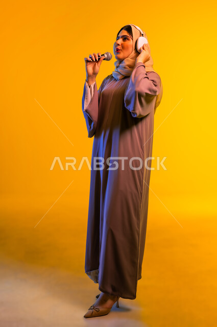 مفهوم الإصغاء واستخدام تقنيات الهيدفون، أجهزة تقنية حديثة، بورتريه لامرأة عربية خليجية عمانية ترتدي الحجاب والعباءة تضع سماعات الرأس تمسك بيدها مكبر الصوت تنظر إلى شيء ما، سيدة سعودية تقوم بإلقاء الخطابات، ممارسة المواهب الفنية، الجسم بالطول الكامل، خلفية صفراء