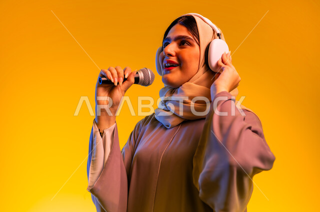 إلقاء الخطابات والمحاضرات، سيدة سعودية تقوم باستخدام تقنيات الهيدفون، الأجهزة التقنية الحديثة، بورتريه مقرب لامرأة عربية خليجية عمانية ترتدي الحجاب والعباءة تضع سماعات الرأس تمسك بيدها مكبر الصوت تنظر إلى شيء ما، ممارسة المواهب الفنية، خلفية صفراء
