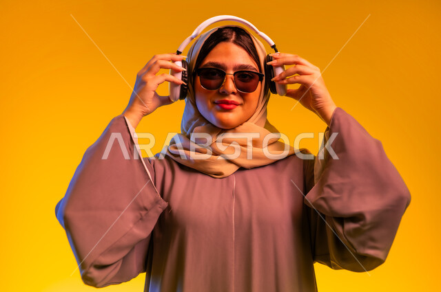 سيدة سعودية تقوم باستخدام تقنيات الهيدفون، قضاء أوقات ممتعة بسماع الأغاني، مشاهدة الأفلام والمسلسلات، بورتريه مقرب لامرأة عربية خليجية عمانية ترتدي الحجاب والعباءة والنظارات الشمسية تضع سماعات الرأس تنظر إلى الكاميرا، الأجهزة التقنية الحديثة، خلفية صفراء