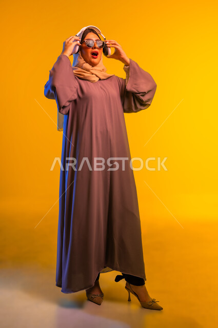 استخدام الأجهزة التقنية الحديثة، سيدة سعودية تقوم بالاستماع إلى الأغاني، مشاهدة الأفلام والمسلسلات، بورتريه لامرأة عربية خليجية عمانية ترتدي الحجاب والعباءة والنظارات الشمسية تضع سماعات الرأس تنظر إلى شيء ما بإيماءات الدهشة، الجسم بالطول الكامل، خلفية صفراء