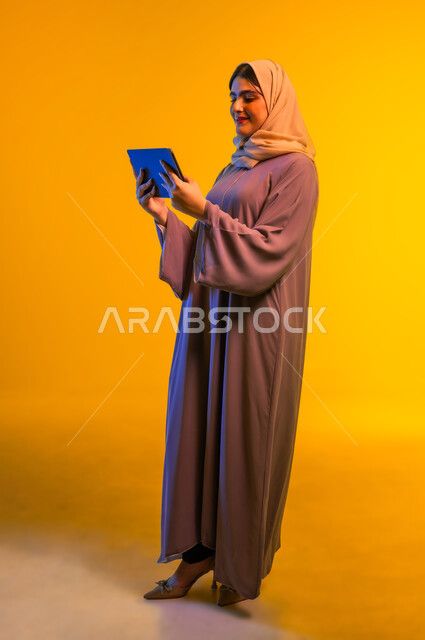 الدردشة مع الأصدقاء وتصفح مواقع التواصل الاجتماعي، استخدام أجهزة تقنية حديثة، بورتريه من الجانب لامرأة عربية خليجية عمانية ترتدي الحجاب والعباءة تقوم بإجراء مكالمة فيديو عبر الجهاز اللوحي، سيدة سعودية تشاهد الأفلام والمسلسلات، الجسم بالطول الكامل، خلفية صفراء