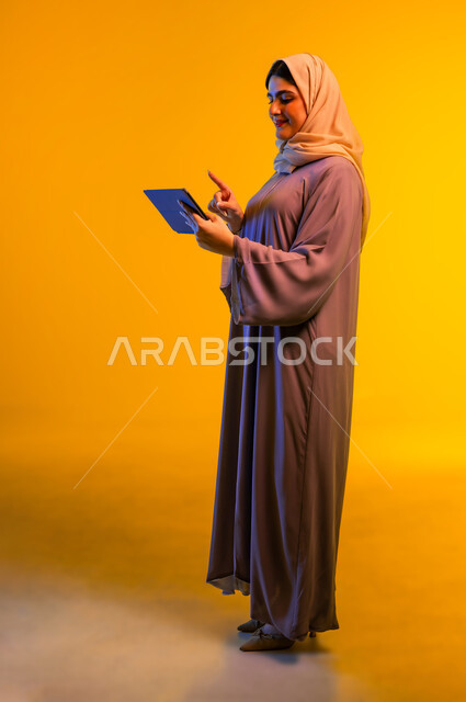 استخدام الأجهزة الحديثة، بورتريه من الجانب لامرأة عربية خليجية عمانية ترتدي الحجاب والعباءة تقوم بالنقر على الجهاز اللوحي، التواصل مع الأهل والأصدقاء، سيدة سعودية تقوم بمشاهدة الأفلام والمسلسلات وتصفح مواقع التواصل الاجتماعي، الجسم بالطول الكامل، خلفية صفراء