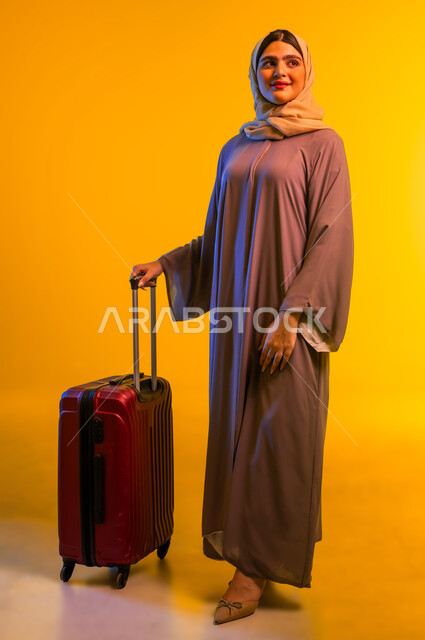 الشعور بالسعادة والفرح، السياحة في إجازة الصيف, شابة سعودية تستعد لقضاء رحلة صيفية ممتعة, بورتريه لشابة عربية خليجية عمانية محجبة ترتدي العباءة تمسك حقيبة السفر بإيماءات الحماس, صورة الجسم بالطول الكامل, خلفية سوداء.