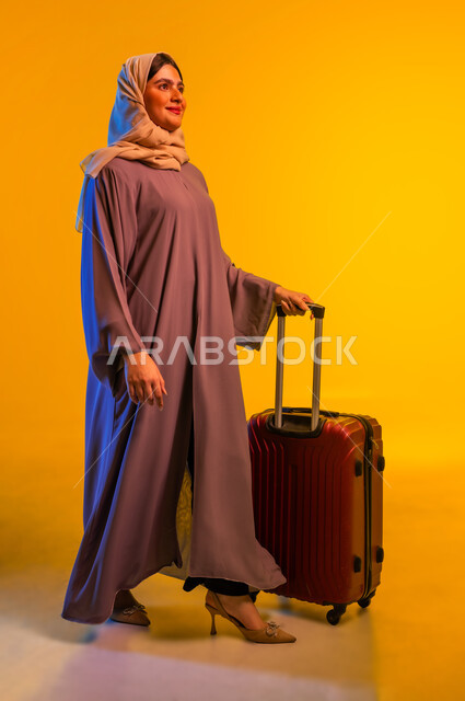 الشعور بالسعادة والفرح، السياحة في إجازة الصيف, شابة سعودية تستعد لقضاء رحلة صيفية ممتعة, بورتريه لشابة عربية خليجية عمانية محجبة ترتدي العباءة تمسك حقيبة السفر بإيماءات الحماس, صورة الجسم بالطول الكامل, خلفية سوداء.