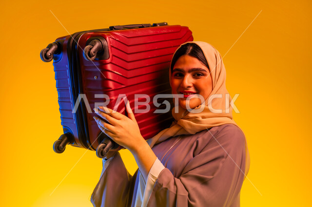 Summer vacation tourism, preparing for a fun and entertaining summer trip, close-up portrait of a veiled young Gulf Omani Arab woman wearing an abaya, carrying a red travel bag on her shoulder, looking at the camera with gestures of enthusiasm and pleasure, yellow background