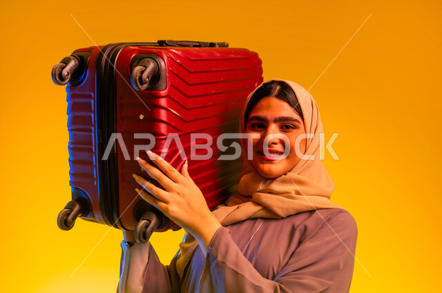 السياحة في إجازة الصيف, الاستعداد لقضاء رحلة صيفية ترفيهية ممتعة, بورتريه مقرب لشابة عربية خليجية عمانية محجبة ترتدي العباءة تحمل حقيبة السفر الحمراءعلى كتفها، شابة سعودية تنظر الى الكاميرا بإيماءات الحماس والسرور, خلفية صفراء