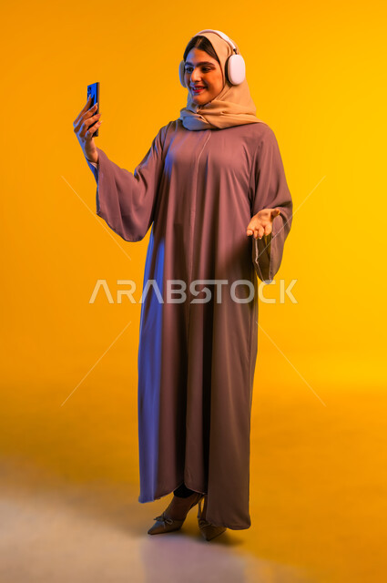 مفهوم الإصغاء والاستمتاع، قضاء وقت ممتع بالاستماع الى الموسيقى, استخدام الهيدفون في اوقات الفراغ، بورتريه مقرب لامرأة عربية خليجية عمانية ترتدي العباءة تضع سماعات الرأس، شابة سعودية تمسك الهاتف المحمول بيدها بإيماءات السعادة، تطور التقنيه والتكنولوجيا، خلفية صفراء