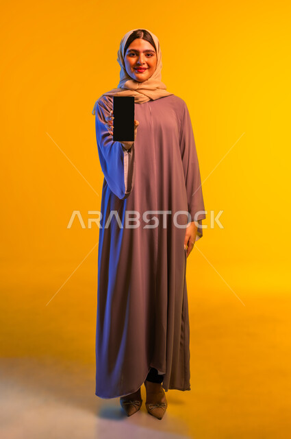 النظر الى الكاميرا بايماءات السعادة والسرور، بورتريه لإمرأة عربية خليجية عمانية مبتسمة ترتدي العباءة تقوم بعرض شاشة سوداء فارغة عبر الهاتف المحمول، شابة سعودية تستخدم الأجهزة والتقنيات الحديثة والمتطورة، الجسم بالطول الكامل، خلفية صفراء