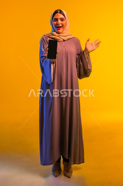 النظر الى الكاميرا بايماءات السعادة والسرور، بورتريه لإمرأة عربية خليجية عمانية مبتسمة ترتدي العباءة تقوم بعرض شاشة سوداء فارغة عبر الهاتف المحمول، شابة سعودية تستخدم الأجهزة والتقنيات الحديثة والمتطورة، الجسم بالطول الكامل، خلفية صفراء