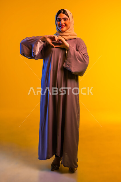 Concept of love and affection, showing feelings of love and passion, heart shaped hand gestures, close-up portrait of veiled Omani Gulf Arab woman wearing abaya looking at camera with expressions of pleasure and happiness, full body image, yellow background