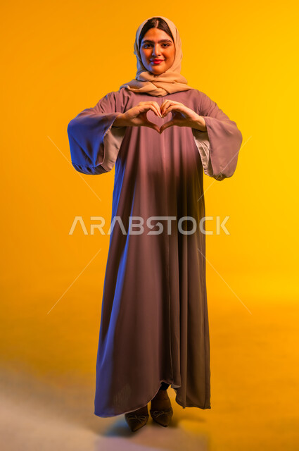 مفهوم المحبة والمودة، شابة سعودية تظهر مشاعر الحب والشغف، ايماءات يدين على شكل قلب، بورتريه مقرب لامرأه عربيه خليجيه عمانية محجبة ترتدي العباءة تنظر إلى الكاميرا بتعابير السرور والسعادة، الجسم بالطول الكامل، خلفية صفراء