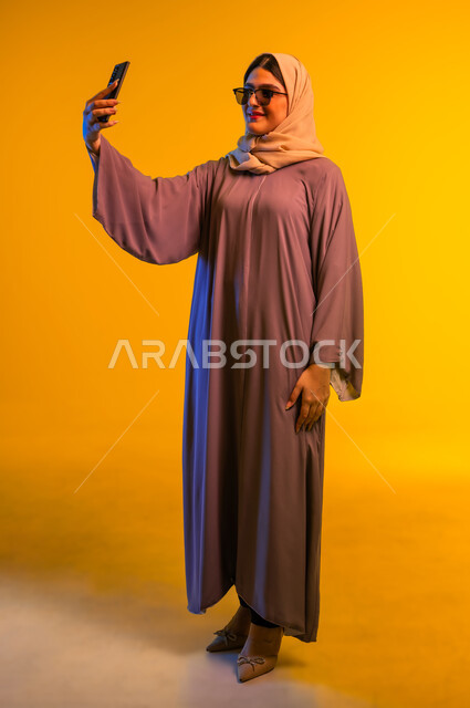 برامج وتطبيقات الجوال المتنوعة، استخدام الأجهزة التقنية الحديثة، شابة سعودية توثق اللحظات السعيدة، بورتريه مقرب لشابة عربية خليجية عمانية محجبة مبتسمة تمسك بيدها جهاز الهاتف المحمول تقوم بالتقاط صور السيلفي التذكارية، الجسم بالطول الكامل، خلفية صفراء